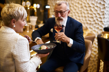 Mature couple of caucasian man and woman sit in restaurant and eat dessert, drink glass of wine. Male in tuxedo, female in white blazer. Woman holding dish
