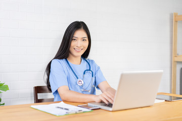 Fototapeta na wymiar Asian female doctor sitting at hospital office desk giving all patient convenience online service advice and smiling write a prescription on laptop to order medical,health care and preventing disease