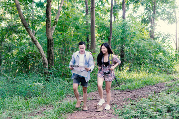 Naklejka na ściany i meble Asian tourist man and woman travel with map in forest. Beautiful Travelers Smiling Couple In Love have a good time in the mountains ,Holding camera In Hands While Traveling Together. Travel Concept