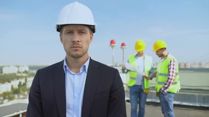 Male architect in helmet looking camera standing rooftop, workers background