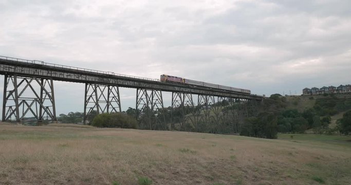 Follow view of a train crossing a old historic narrow trestle railway bridge from a field below the bridge