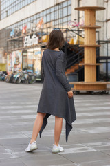 Full length outdoor portrait. Woman in gray dress and white sneakers walking urban city street. Caucasian female model with long brunette wavy hair