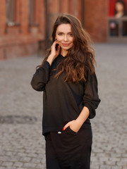 Fototapeta premium Outdoor summer portrait. Young elegant woman in black hoodie and trousers standing and posing at urban city street