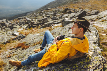 relaxes young man in fashion yellow raincoat, green hat exploring landscape, side view photo. relaxation, pastime
