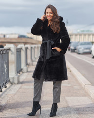 Obraz premium Outdoor portrait of beautiful young pretty woman in black vegan and faux fur coat standing and posing at urban city embankment. Fashion trendy style clothes