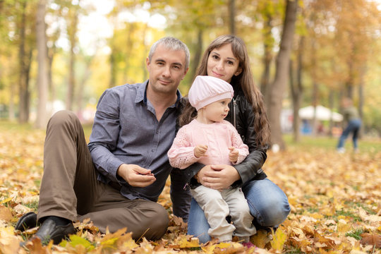 Happy married couple sitting on leaves in autumn and holding a beautiful baby daughter .parents kiss the child sitting on the autumn leaves