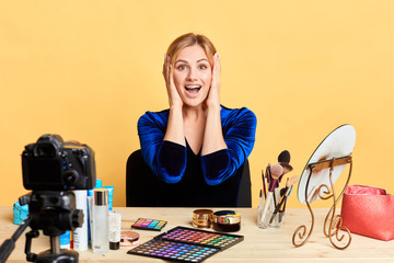 Front view of impressed pretty blonde blogger sits in makeup room with palms near her face, expresses happiness and amazement, juts received great news. Massmedia and positive emotions concept.