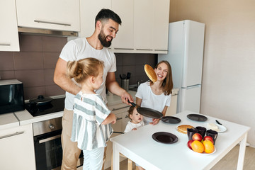 cheerful young father cooking pancakes, holding pan, throwing pancakes over it while wife and his adorable daughters watching him in the kitchen. lifestyle, free time, spare time