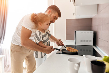 young father and his beautiful blonde daughter enjoying cooking pancakes for mother in the kitchen at home - 294132229