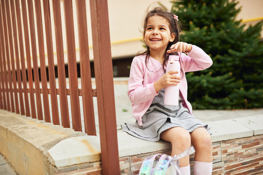 Outdoors image of cute little girl preschooler drinking water from her eco glass bottle sitting against buildings. Happy kid pupil relaxing outside after preschool lessons. People, education concept