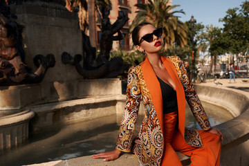 Fashion style outdoor portrait. Elegant woman in colorful orange costume, sunglasses and black shoes sitting on stone fence of fountain on a sunny evening in tropical city