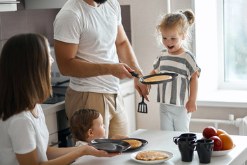 man in white T-shirt serving pancakes to family, father holding a pan with hot pancake, close up...