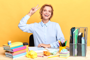 funny woman in shirt showing two fingers while sitting at the table with documents and sheets of paper. close up portrait, body language , isolated yellow background, studio shot