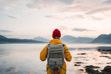Back view of male tourist with rucksack standing on coast in front of great mountain massif while journey.  Man traveler wearing yellow jacket with backpack explore nature. Wanderlust lifestyle - obrazy, fototapety, plakaty