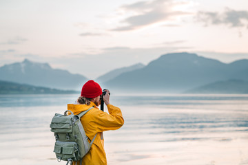 Traveler photographer wearing yellow raincoat taking photo by professional camera of fantastic scandinavia nature. Man tourist tourist travels to beautiful mountain and fjord places - obrazy, fototapety, plakaty