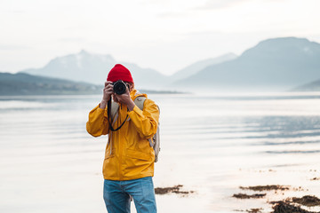 Hipster traveler photographer takes a photo beautiful nordic landscape on professional camera. Man tourist with backpack shoots a photo scandinavia nature - obrazy, fototapety, plakaty