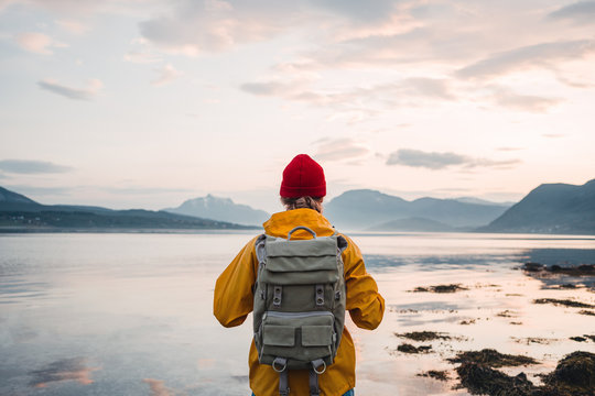 Back view of male tourist with rucksack standing on coast in front of great mountain massif while journey.  Man traveler wearing yellow jacket with backpack explore nature. Wanderlust lifestyle