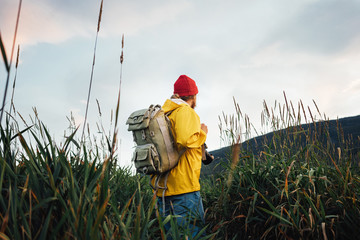 Hipster traveler wearing backpack and yellow raincoat looking away at mountain. Alone man traveling at scandinavian authentic landscape