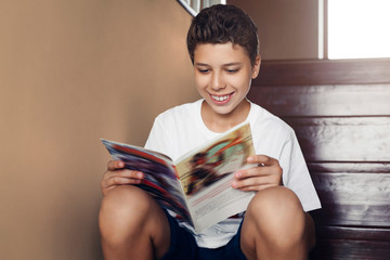 Smiling teen boy sitting at home on steps, reading magazine. Cheerful guy in white t-shirt is...