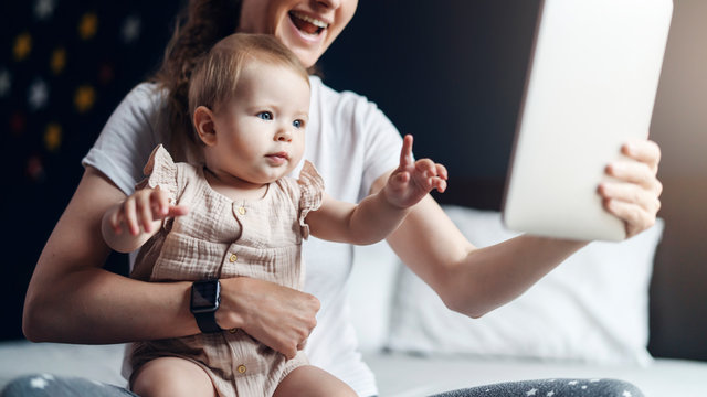 Nine-month-old Baby Girl With Mom Sitting On Bed, Looking At Screen Of Digital Tablet. Child Development Using Modern Technology.