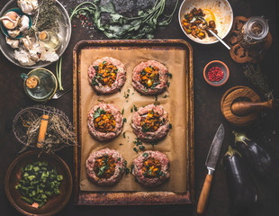 Preparation of minced meat patties in bagel shape filled with roasted vegetables on baking sheet on rustic kitchen table with herbs, spices and utensils. Top view. Low carb food concept