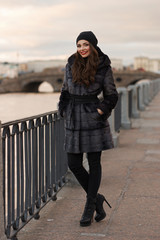 Beautiful lady in gray fur coat and cashmere cap at river embankment on a cold day. Fall winter fashion look.