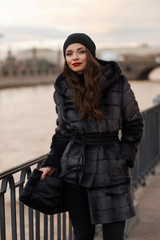 Obraz premium Beautiful lady in gray fur coat and cashmere cap at river embankment on a cold day. Fall winter fashion look.