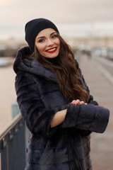 Obraz premium Beautiful lady in gray fur coat and cashmere cap at river embankment on a cold day. Fall winter fashion look.