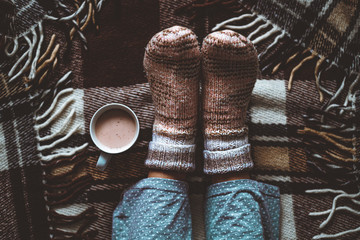 Cozy woman in knitted winter warm socks and in pajamas resting on checkered plaid blanket with a cup of hot cocoa at home in winter time. Cozy time and winter drinks. Top view