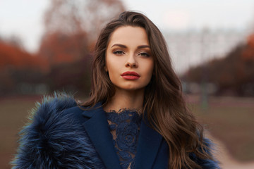 Calm young fashionable woman in blue coat and fur scarf posing in autumn park. Fall fashion look....