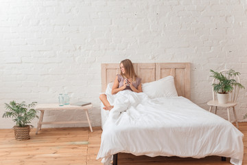 Pretty smiling girl sitting in her bed and drinking morning tea, bedroom in loft style