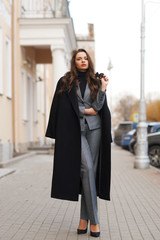 Fall fashion look. Stylish woman in gray suit and black coat standing alone at city street in old town and looking in camera. Elegant lady full length portrait