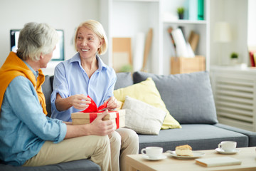 Portrait of excited senior woman opening gift from caring husband sitting on comfortable sofa in home interior, copy space