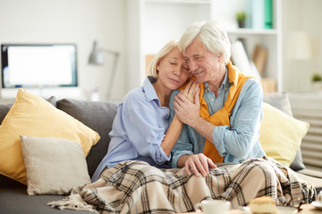 Portrait of loving senior couple embracing tenderly sitting on comfortable couch at home, covered...