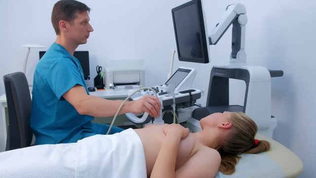 Man doctor making ultrasound diagnostic of mammary glands of young woman on bust. He runs ultrasound sensor over patient's mammary glands and looks at image on screen. Diagnosis of internal organs.