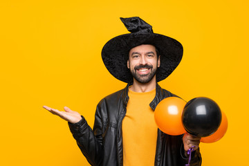Man with witch hat holding black and orange air balloons for halloween party holding copyspace imaginary on the palm to insert an ad