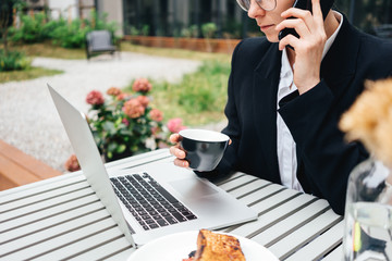 Young businesswoman sitting at cafe on veranda having telephonic conversation. Beautiful female business talking on smartphone while sitting at coffee shop and working on laptop
