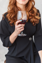 Woman with long red hair in black dress holds a glass with red wine in hands