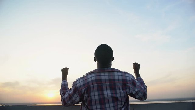 Back view of black man raising hands to sky against sunset, achievement, winner
