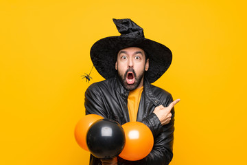 Man with witch hat holding black and orange air balloons for halloween party surprised and pointing side