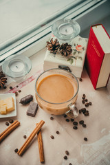 Fototapeta na wymiar Cup of coffee with milk near the window, book, cinnamon sticks, cheese, candy, cones, candles