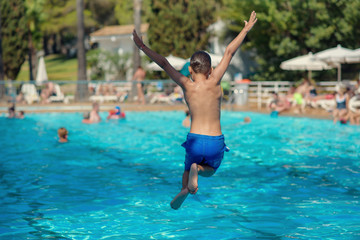 Cute active European boy in blue swimming shorts having fun in hotel’s swimming pool, he is making fantastic jump, his hands open wide.