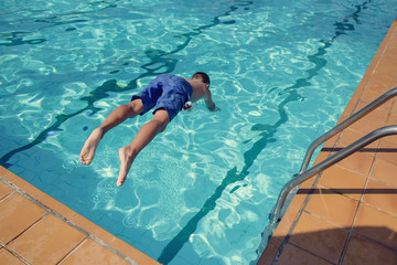 Cute active European boy in blue swimming shorts having fun in hotel’s swimming pool, he is making fantastic jump, back view.