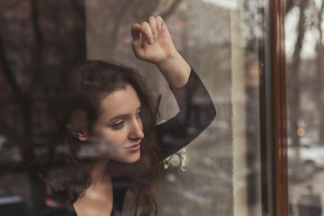 Fototapeta na wymiar Attractive girl with long brunette hair behind the glass, coffee shop window, concept of female emotions