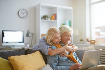 Portrait of senior man using laptop while working from home with loving wife embracing him, copy...