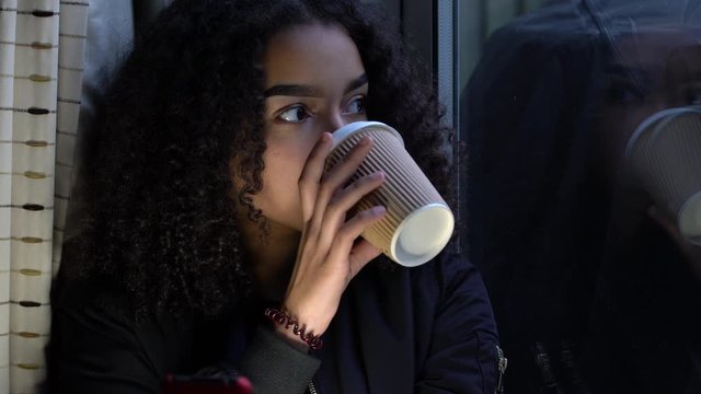 Sad depressed or thoughtful beautiful female mixed race African American girl teenager young woman teen sitting using her mobile cell phone looking out of a window and drinking cup of coffee