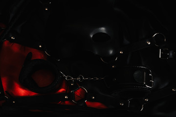 Top view of bdsm leather kit (whips, handcuffs, mask, anal plug) against of a black and a red silk.