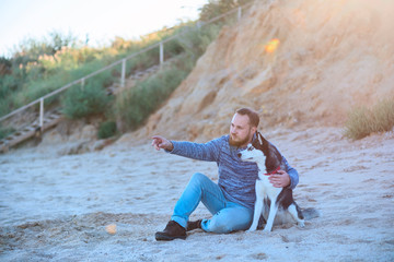 Bearded man in jeans and sweater sits on the sand by the sea with his dog Siberian husky breed