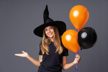 Young witch holding black and orange air balloons extending hands to the side for inviting to come