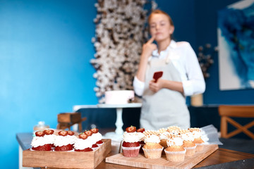 Thoughtful young woman touching her chin with finger, thinking about the decoration of muffins, blurred background, studio shot.